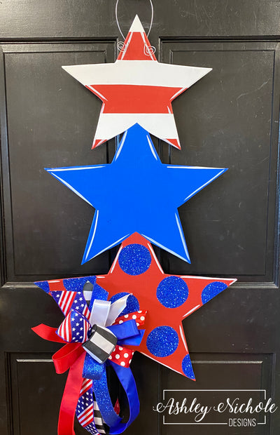 A patriotic door hanger featuring layered star shapes in red, white, and blue colors with decorative elements and text that says 'Ashley Nichole Designs'.